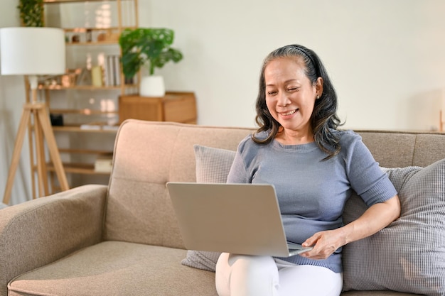 Alegre mulher aposentada de idade asiática gosta de assistir a filmes em seu laptop em sua sala de estar