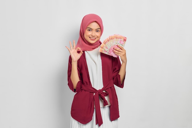 Alegre mulher asiática bonita em camisa casual e hijab segurando notas de rupia indonésias, mostrando o gesto okey isolado sobre fundo branco. Conceito de estilo de vida religioso de pessoas