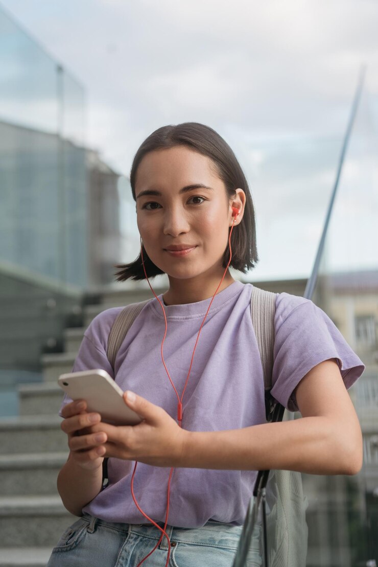Foto alegre mulher asiática vestindo camiseta roxa segurando celular ouvindo música olhando para a câmera