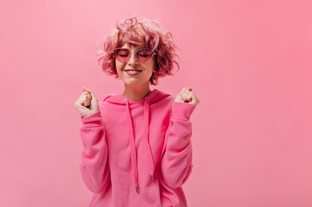 Foto alegre mulher bonita com capuz rosa brilhante faz desejo e posa com olhos vestidos garota de cabelo rosa em poses de óculos de sol em fundo isolado