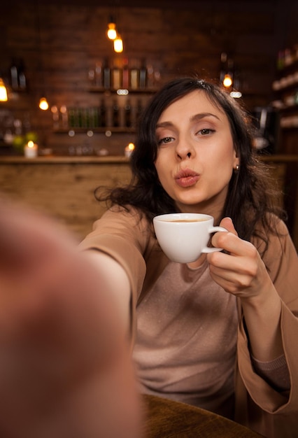 Alegre mulher bonita dando beijos e tirando um auto-retrato com smartphone em uma cafeteria. Menina segurando uma xícara de café. Cafeteria moderna.