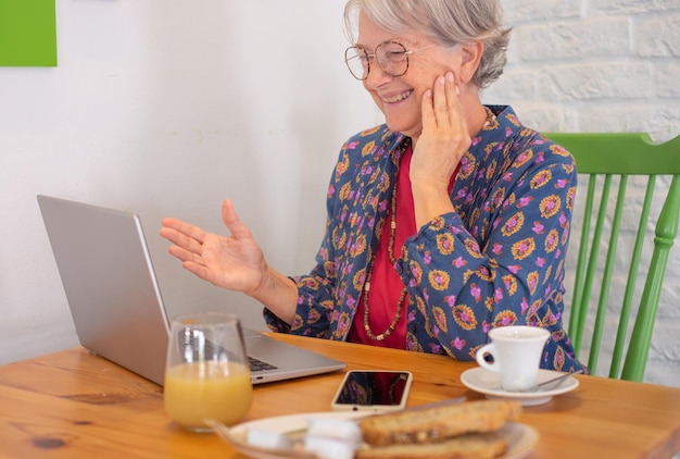Alegre mulher caucasiana sênior sentada à mesa do café no café da manhã usando laptop no bate-papo por vídeo Senhora idosa de cabelos grisalhos sorrindo feliz desfrutando de tecnologia e social