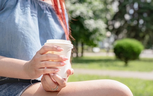 Alegre mulher da moda em roupas de verão bebendo café no parque com a mão fechada com xícara