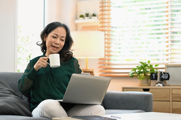 Alegre mulher de meia idade em roupas casuais bebendo café e usando um laptop no sofá em casa