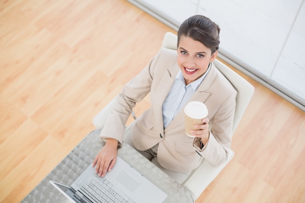 Alegre mulher de negócios de cabelos castanhos inteligente segurando uma xícara de café enquanto usa um laptop