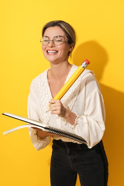 Alegre mulher de óculos segurando grande lápis e caderno em fundo amarelo