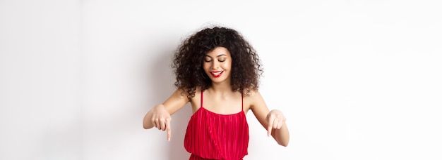 Alegre mulher elegante em vestido vermelho e maquiagem olhando e apontando para baixo com um sorriso satisfeito mostrando