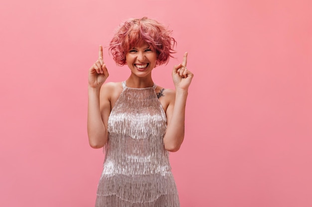 Alegre mulher encaracolada de vestido prateado sorri amplamente e aponta os dedos para cima no lugar para texto em fundo rosa isolado