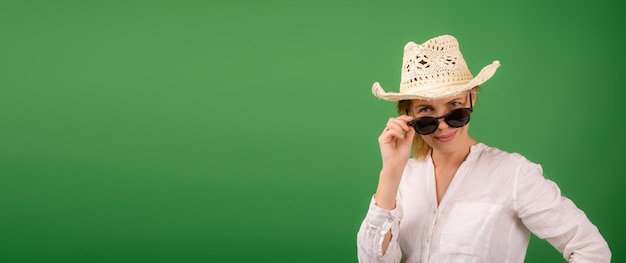 Alegre mulher espantada em um chapéu e óculos de sol em uma camisa branca sobre um fundo verde Olha para a câmera com os óculos para baixo Lugar de banner para texto