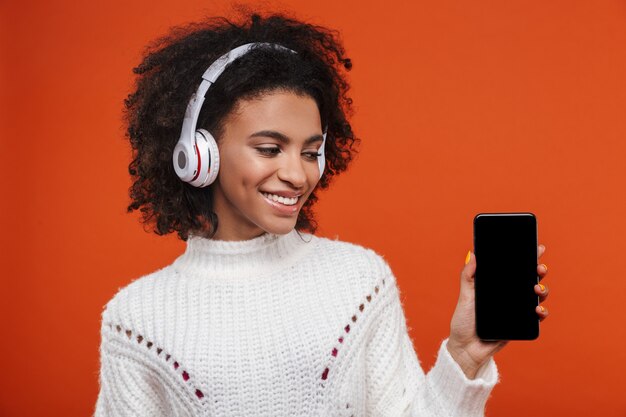 Alegre mulher jovem e atraente africana usando fones de ouvido sem fio, isolado na parede vermelha, mostrando a tela em branco do celular