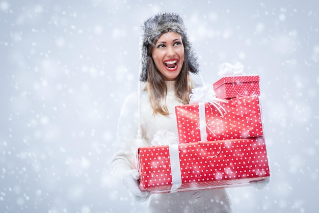 Alegre mulher jovem e bonita em agasalhos segurando muitos presentes de Natal enquanto nevando.