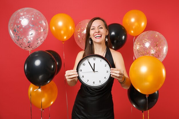 Alegre mulher jovem e bonita em vestido preto, comemorando, segurando o relógio redondo em balão de ar de fundo vermelho. Dia dos namorados, dia internacional da mulher, feliz ano novo aniversário maquete feriado conceito festa.