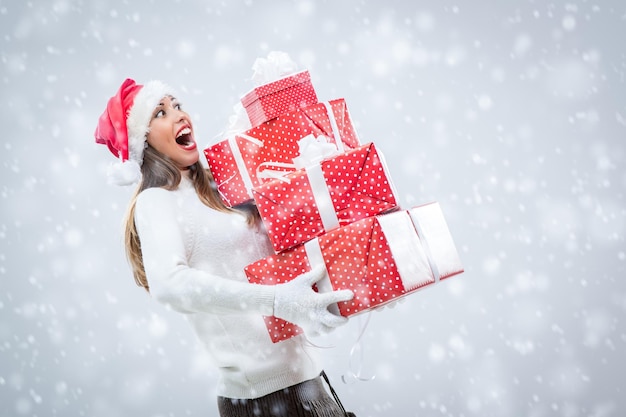 Alegre mulher jovem e bonita usando chapéu de Papai Noel e segurando muitos presentes de Natal enquanto neva.