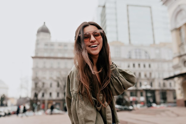 Alegre mulher morena com longos cabelos voadores em óculos castanhos e jaqueta verde sorri sinceramente ao ar livre Senhora feliz ri e anda de bom humor lá fora