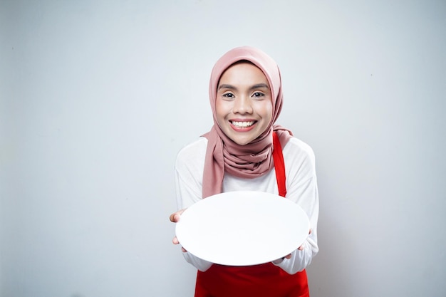 Alegre mulher muçulmana asiática em avental vermelho segurando prato vazio Anúncio de comida Conceito de culinária