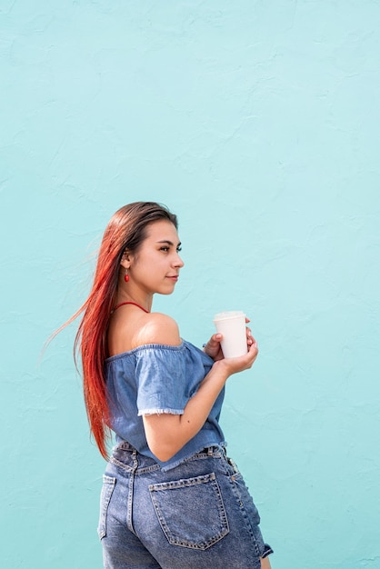 Alegre mulher na moda com cabelo vermelho bebendo café dançando no fundo da parede azul na rua