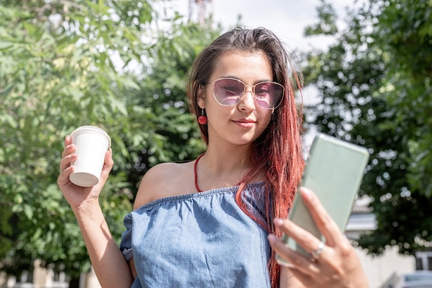 Alegre mulher na moda com cabelo vermelho tomando café no parque tomando selfie