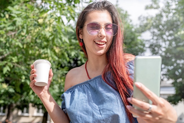 Alegre mulher na moda com cabelo vermelho tomando café no parque tomando selfie