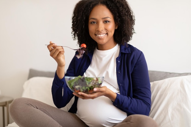 Foto alegre mulher negra milenar encaracolada em roupas domésticas com barriga grande gosta de gravidez come salada