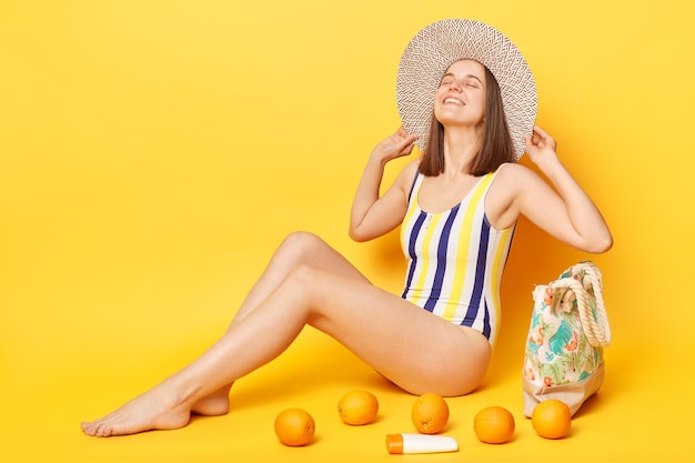 Alegre mulher satisfeita vestindo maiô listrado e chapéu isolado em fundo amarelo tomando banho de sol na praia expressando felicidade parece encantada