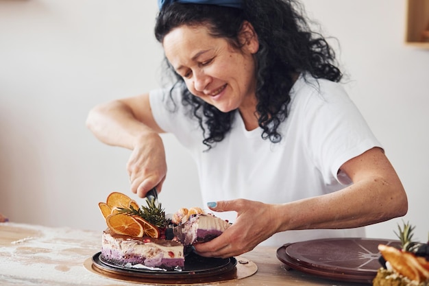 Alegre mulher sênior com cabelo preto encaracolado fazendo fatias de bolo dietético fresco na cozinha