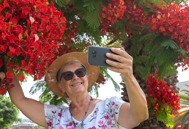 Alegre mulher sênior com chapéu de palha tomando selfie em um agradável parque público com plantas de flores vermelhas bonitas e conceito de palmeiras de idosos ativos nas férias de verão