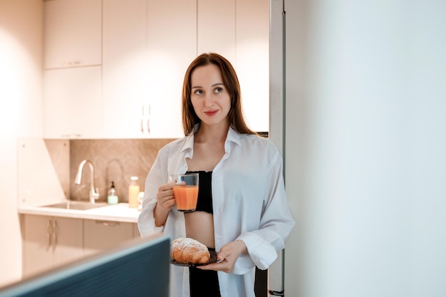 Alegre mulher sorridente bebendo suco e comendo croissant na cozinha de casa Conceito de estilo de vida saudável