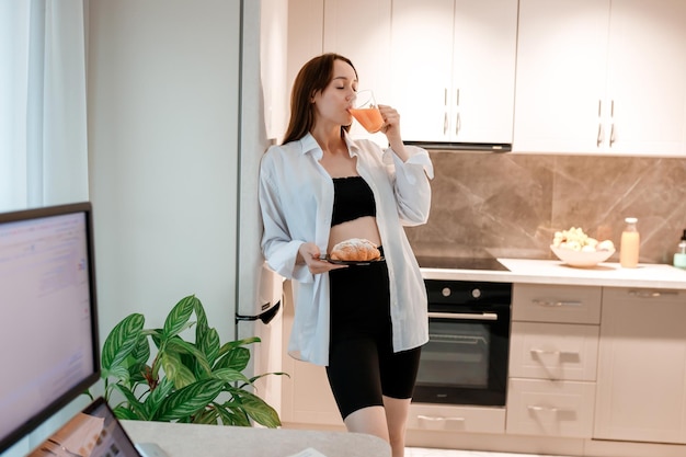 Alegre mulher sorridente bebendo suco e comendo croissant na cozinha de casa Conceito de estilo de vida saudável