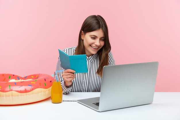 Alegre mulher sorridente inserindo dados de passaporte no laptop, reservando ingressos e férias on-line, preparando-se para feriados, anel de borracha e limonada na mesa. Tiro de estúdio interior isolado no fundo rosa