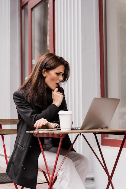 Alegre mulher surpresa sorrindo e olhando para a quantidade de ganhos no cassino online e se regozijando do lado de fora Garota chocada está ganhando uma aposta no cassino online e sentada com o laptop no café ao ar livre