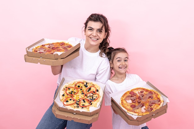 Alegre niña adulta y su hermana pequeña con pizza en cajas para entrega sobre un fondo rosa.