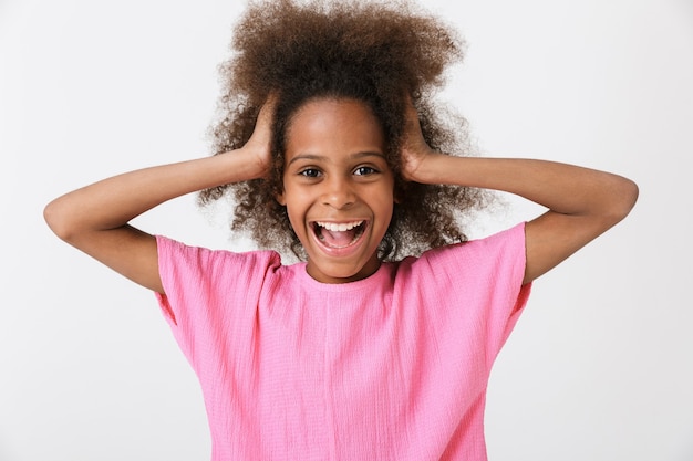 Alegre niña africana divertida vistiendo blusa rosa que se encuentran aisladas sobre la pared blanca, haciendo muecas, gritando