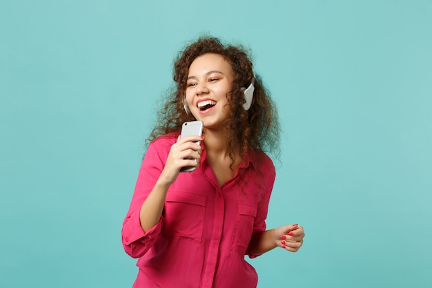 Alegre niña africana en ropa casual mantenga teléfono móvil, escuchando música con auriculares aislados sobre fondo azul turquesa en estudio. Concepto de estilo de vida de emociones sinceras de personas. Simulacros de espacio de copia.