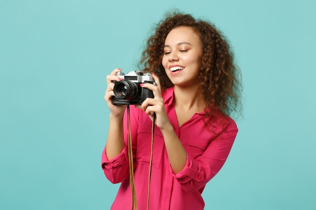 Alegre niña africana en ropa casual tomando fotografías en una cámara de fotos vintage retro aislada sobre fondo de pared azul turquesa en estudio. Concepto de estilo de vida de emociones sinceras de personas. Simulacros de espacio de copia.