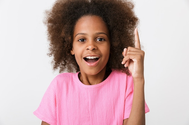 Alegre niña africana vistiendo blusa rosa que se encuentran aisladas sobre la pared blanca, señalando con el dedo en el espacio de copia