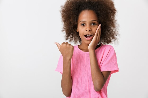 Alegre niña africana vistiendo blusa rosa que se encuentran aisladas sobre la pared blanca, señalando con el dedo en el espacio de copia