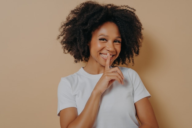 Alegre niña afro sonriente mostrando el signo de shhh, guardar gesto de silencio, con el dedo índice cerca de los labios, de pie sobre un fondo beige pastel y mirando a la cámara. Emociones positivas y lenguaje corporal.