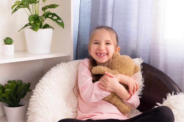 Una alegre niña de años con una camiseta rosa sentada en un sillón en casa abrazando un oso de peluche