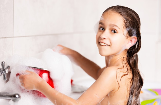 Alegre niña caucásica juega con espuma mientras se baña en la bañera