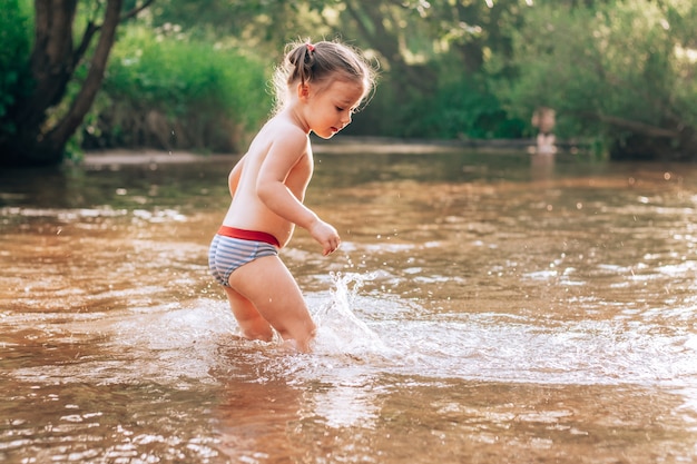 Alegre niña divertida 3-4 se encuentra en el río, salpicaduras de rayos de sol