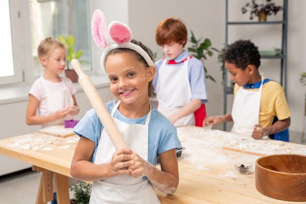 Alegre niña rizada en delantal cruzando los brazos sobre el pecho mientras está de pie junto a la mesa de la cocina con ingredientes para galletas