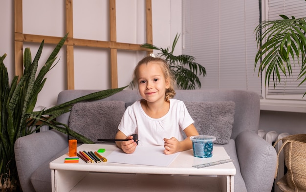 Una alegre niña rubia está sentada en una mesa y dibuja con lápices sobre papel