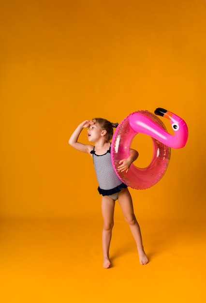 Alegre niña rubia en un traje de baño a rayas sostiene un círculo inflable rosa y mira hacia otro lado en una superficie amarilla con un lugar para el texto