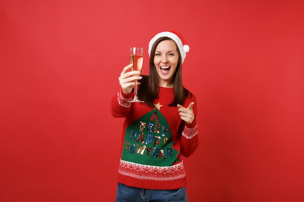 Alegre niña de Santa con sombrero de Navidad apuntando con el dedo índice a la cámara, sosteniendo una copa de champán aislado sobre fondo rojo. Feliz año nuevo 2019 celebración concepto de fiesta navideña. Simulacros de espacio de copia.