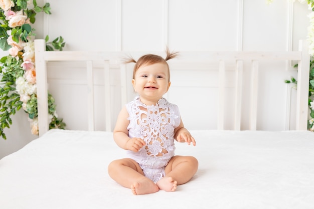 Alegre niña de seis meses sentada en una hermosa habitación luminosa en una cama blanca