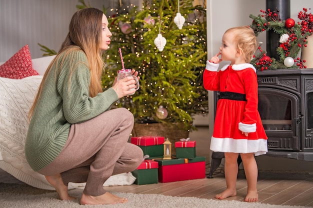 Alegre niño feliz con traje rojo de Papá Noel con mamá en un dormitorio decorado celebrando las vacaciones de Navidad en casa