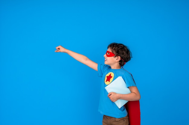 Alegre niño vestido con traje de superhéroe sostenga el libro y extiende su mano