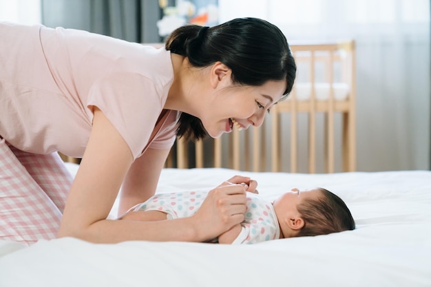 Alegre nova mãe coreana ajoelhada na cama e dizendo bom dia para seu bebê. vista lateral da mulher olhando e conversando com seu lindo filho no quarto.