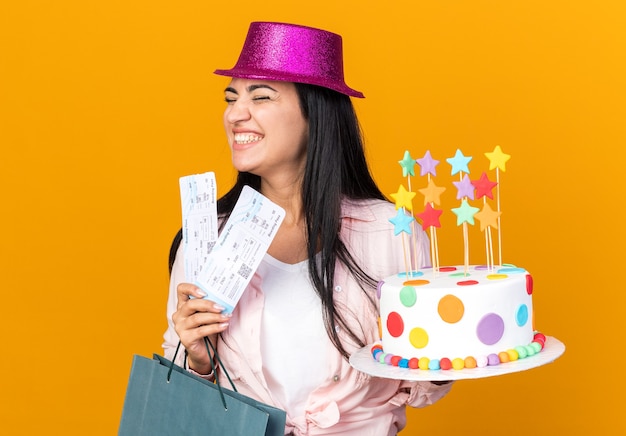 Alegre con los ojos cerrados hermosa joven vistiendo gorro de fiesta sosteniendo una bolsa de regalo con pastel y boletos