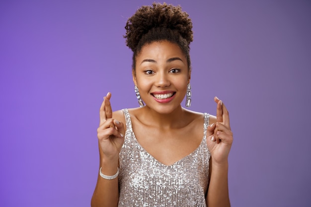 Alegre, optimista, encantadora novia afroamericana que apoya a un amigo que desea ganar el primer premio dedos cruzados suerte sonriendo ampliamente usar un vestido plateado brillante anticipando la buena fortuna.